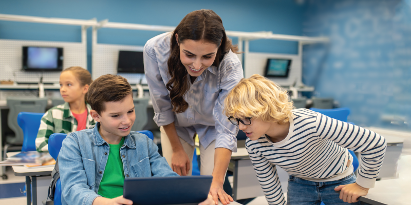 A teacher helps two young students using a tablet in a tech-enhanced classroom, demonstrating effective use of technology in education